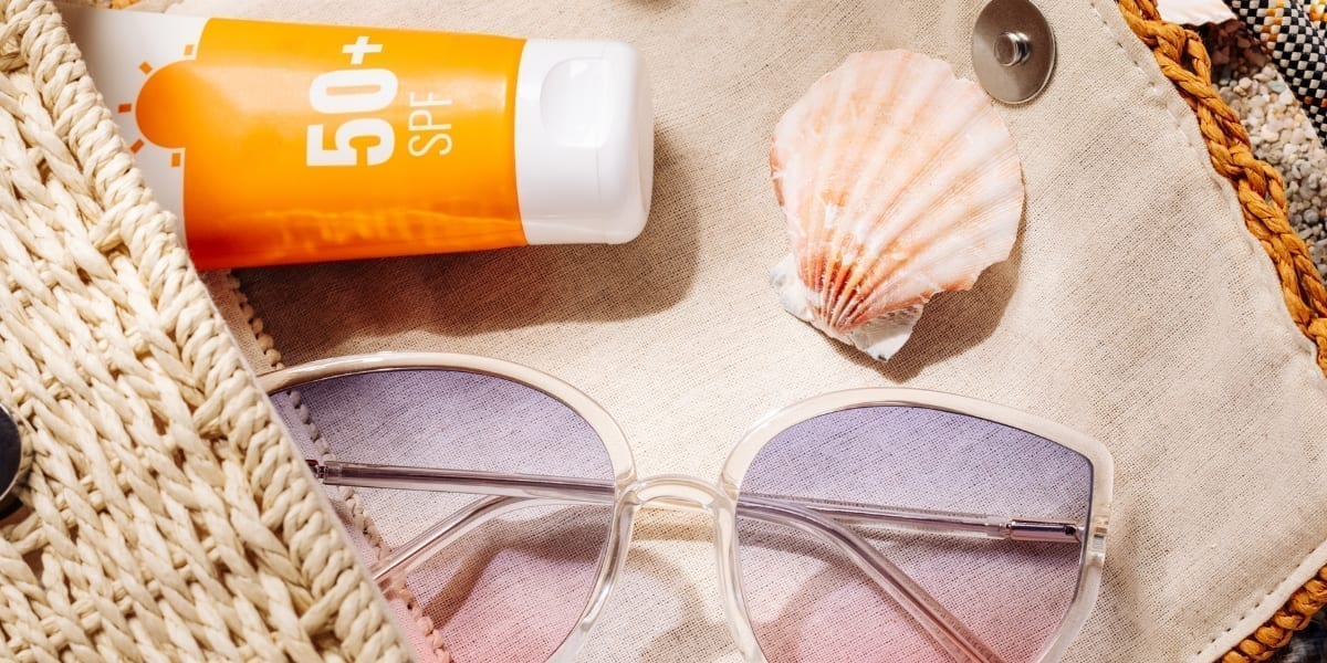 Produse de beauty esențiale pentru vară așezate pe nisip: cremă cu SPF 50, apă termală, balsam de buze și gel cu aloe vera